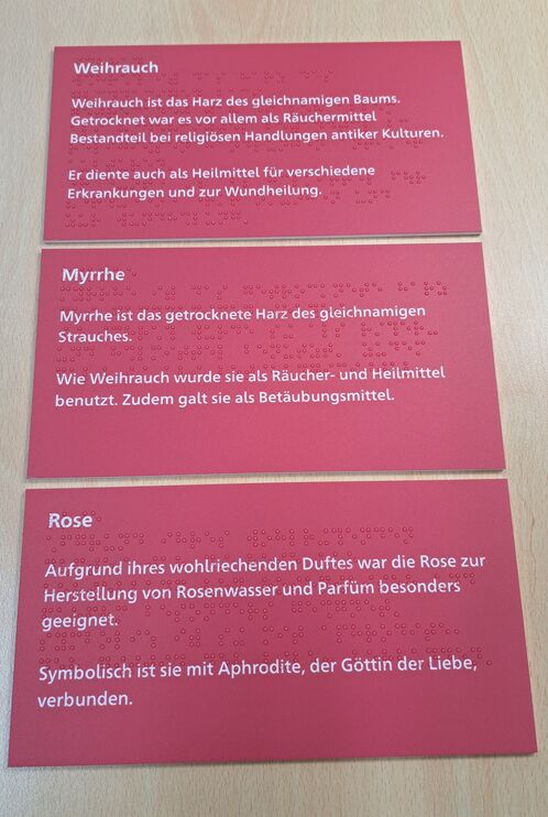 Drei rechteckige, rote Acrylschilder mit weißer Schrift und Braillelack