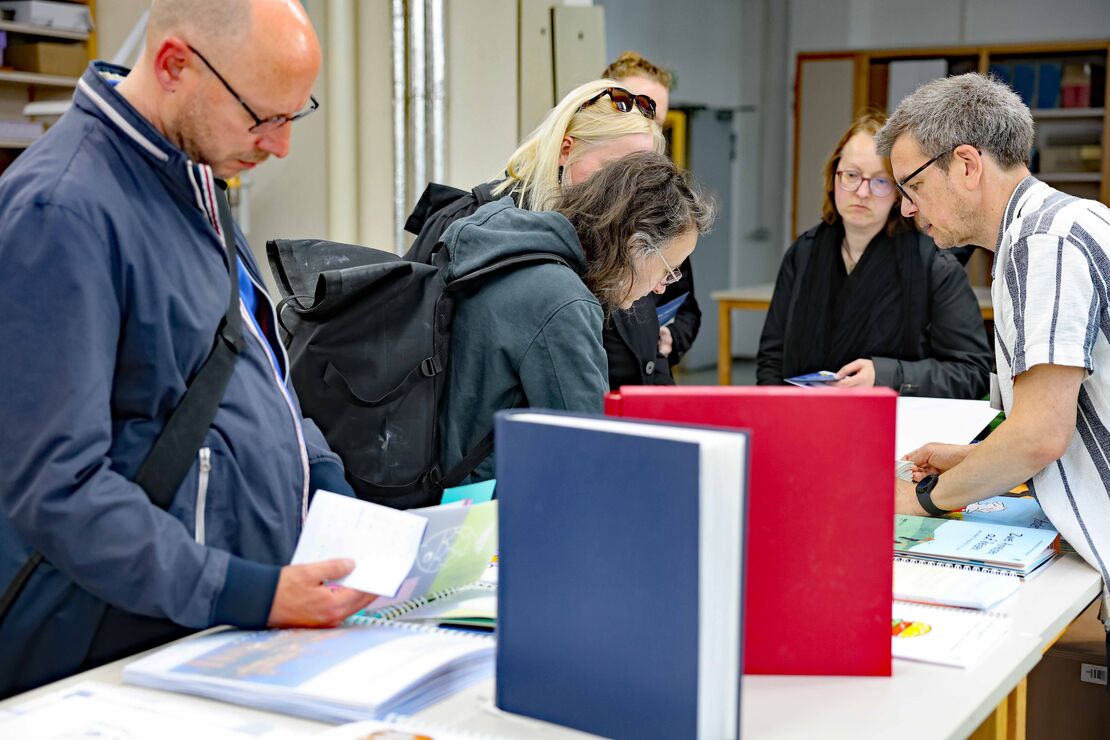 Mehrere Personen schauen sich auf dem Tisch ausliegende Reliefbücher an. Rechts erklärt ein Mann mit Brille den Personen etwas.