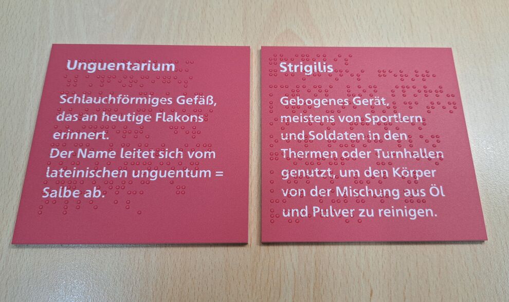 zwei rechteckige, rote Schilder mit Text in weißer Schrift und mit Braillelack