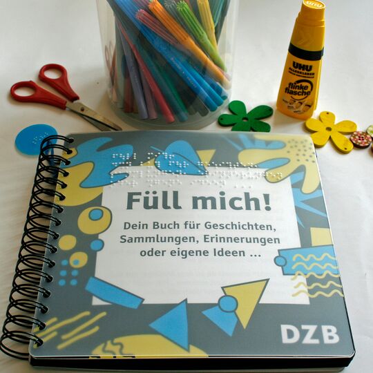 Cover des Ringbuches "Füll mich", oben eine Schere, Filzstifte, Klebstift und kleine, bunte Holzblumen und Holzknöpfe