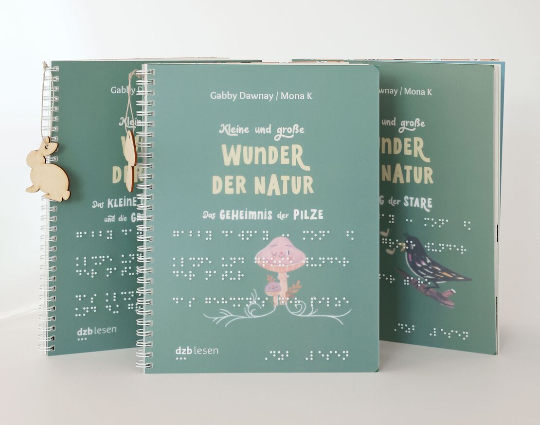 Drei stehende Ringbücher mit dem Titel "Wunder der Natur" in Braille- und Großschrift, im Vordergrund das Cover mit einem Pilz "Das Geheimnis der Pilze"