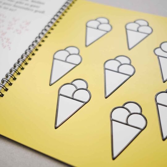 Aufgeschlagenes Ringheft "Mein erster Rätselspaß" mit dem Motiv acht mal Eistüte mit Kugeln, links angedeutet Seite mit Braille- und Großschrift