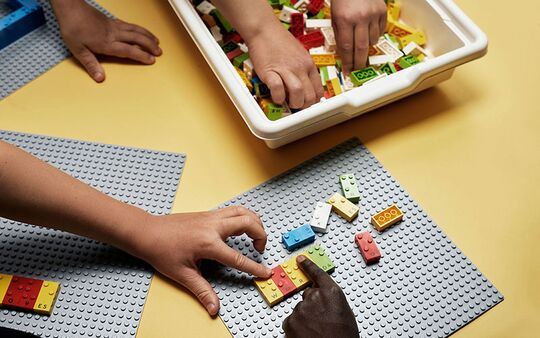 Eine dunkle und eine weiße Hand zeigen auf Braille-Steine, oben zwei Hände greifen sich aus Schale Braille-Steine