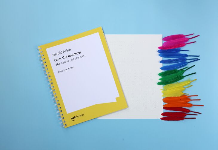 Ringbuch "Harold Asten: Over the Rainbow" mit Braillenotenschrift, rechts daneben ein einzelnes Notenblatt in Braille mit am rechten Rand aufgereihten Federn in Regenbogefarben.