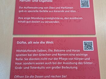 zwei rechteckige, rote Schilder mit Text in weißer Schrift, mit Braillelack und QR-Code