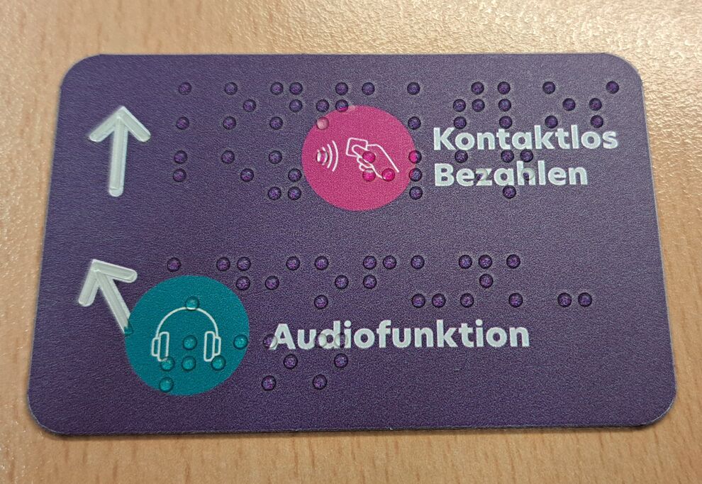 rechteckiges, lilafarbendes Schild mit Symbolen Kopfhörer und Hand mit EC-Karte, Text: Audiofunktion und Kontaktlos bezahlen in weißer Schrift und Brailleschrift