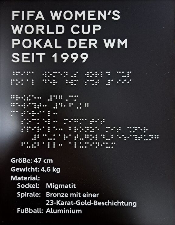schwarzes, rechteckiges Schild mit weißer Profilschrift und Brailleschrift: FIFA Women's World Cup Pokal der WM seit 1999