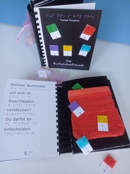 Aufgeschlagenes Ringbuch (linke Seite Text in Braille- und Großschrift, rechte Seite ein Waschlappen aus Stoff und kleine tastbare, rechteckige Pappstücke mit Buchstaben in Braille, über dem Buch stehe das Gleiche