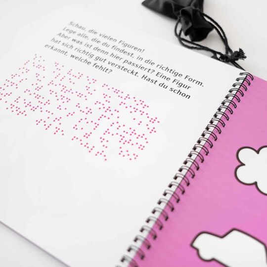 Aufgeschlagenes Ringheft "Mein erster Rätselspaß" mit Motiven Blume, Apfel Auto rechts, links Seite mit Braille- und Großschrift