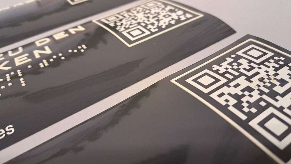 Detailansicht eines Aufklebeschildes mit taktiler Profilschrift, Brailleschrift und QR-Code