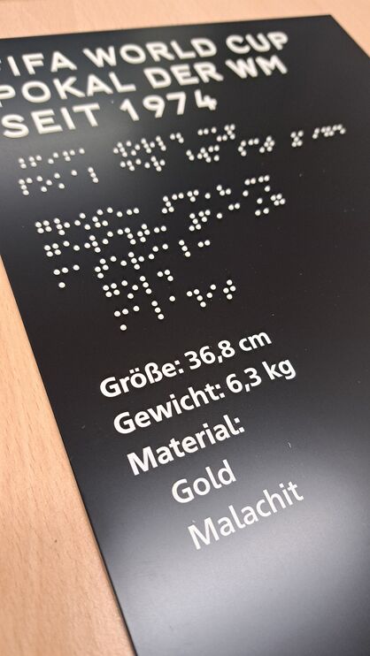 schwarzes, rechteckiges Schild mit weißer Profilschrift und Brailleschrift