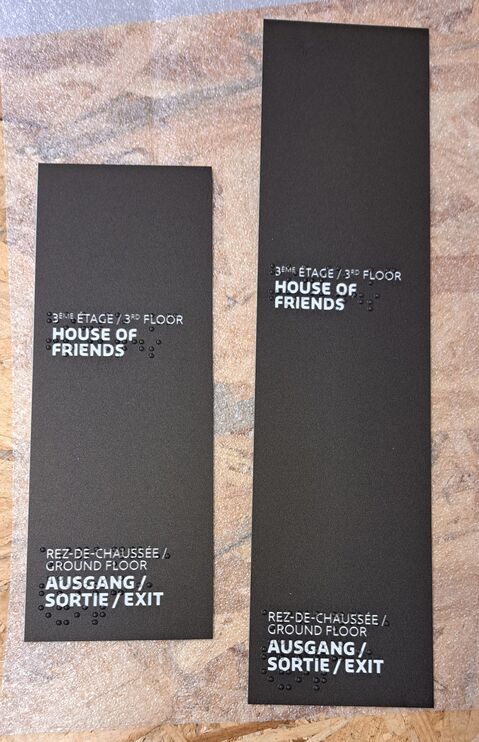 zwei rechteckige, schwarze Schilder im Hochformat mit Text in Brailleschrift und weißer Schrift