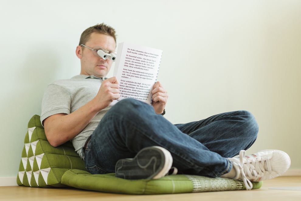 Junger Mann mit Vergrößerungsgläsern sitzt auf Kissen und hält ein Buch in den Händen