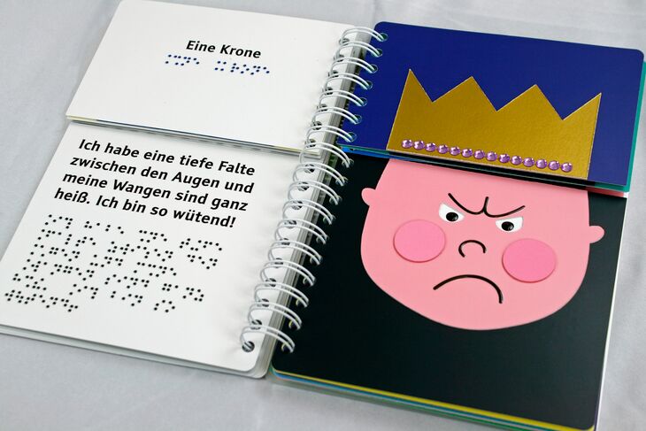 Aufgesschlagenes Ringbuch (links Braille- und Großdruck-Text, rechts: Gesicht eines erzürnten Jungen mit zusammengezogener Stirn und goldener Krone)