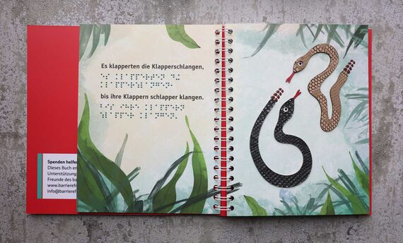 Aufgeschlagenes Ringbuch vor graumeliertem Hintergrund, Doppelseite: links Text in Braille- und Schwarzschrift, rechst eine taktile Illustration (zwei Schlangen und Gras)