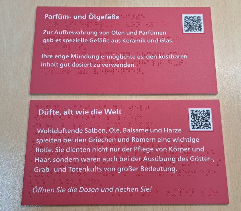 zwei rechteckige, rote Schilder mit Text in weißer Schrift, mit Braillelack und QR-Code