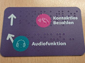rechteckiges, lilafarbendes Schild mit Symbolen Kopfhörer und Hand mit EC-Karte, Text: Audiofunktion und Kontaktlos bezahlen in weißer Schrift und Brailleschrift